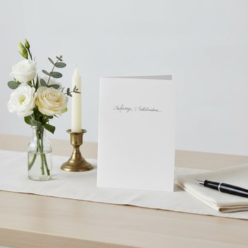 Preview: Trixi Gronau sympathy card AUFRICHTIGE ANTEILNAHME, B6 portrait in pure white with delicate grey calligraphy. Standing on a quiet light desk with cream linen runner, beside a small still bouquet of white Eustoma, a cream rose and eucalyptus in a simple gl