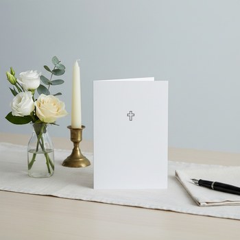 Preview: Trixi Gronau sympathy card KREUZ, B6 portrait in pure white with a fine dark-grey outline cross centered in the upper middle. On a light desk with cream linen runner, beside white Eustoma, cream rose and eucalyptus in a glass vase, a slim cream taper in a