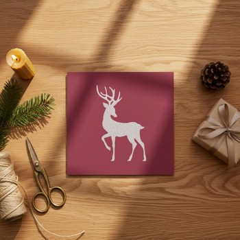 Preview: Trixi Gronau Weihnachtskarte GLITTER HIRSCH, quadratisches Format in tiefem Weinrot mit silbern glitzernder Hirsch-Silhouette. Liegt auf einem warmen Eichen-Schreibtisch zwischen Tannenzweig, Bienenwachskerze, antiker Messing-Schere, Jute-Garn-Rolle, kraf