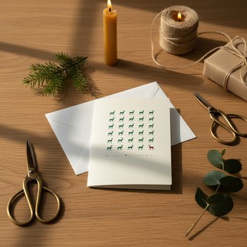Preview: Trixi Gronau Weihnachtskarte MINI REHREIHE, cremefarbene Klappkarte mit Raster aus dunkelgruenen Reh-Silhouetten (eins in Rot) und Frohe Weihnachten in feiner Kalligraphie, dahinter passender Umschlag. Auf warmem Eichen-Schreibtisch mit Tannenzweig, Biene