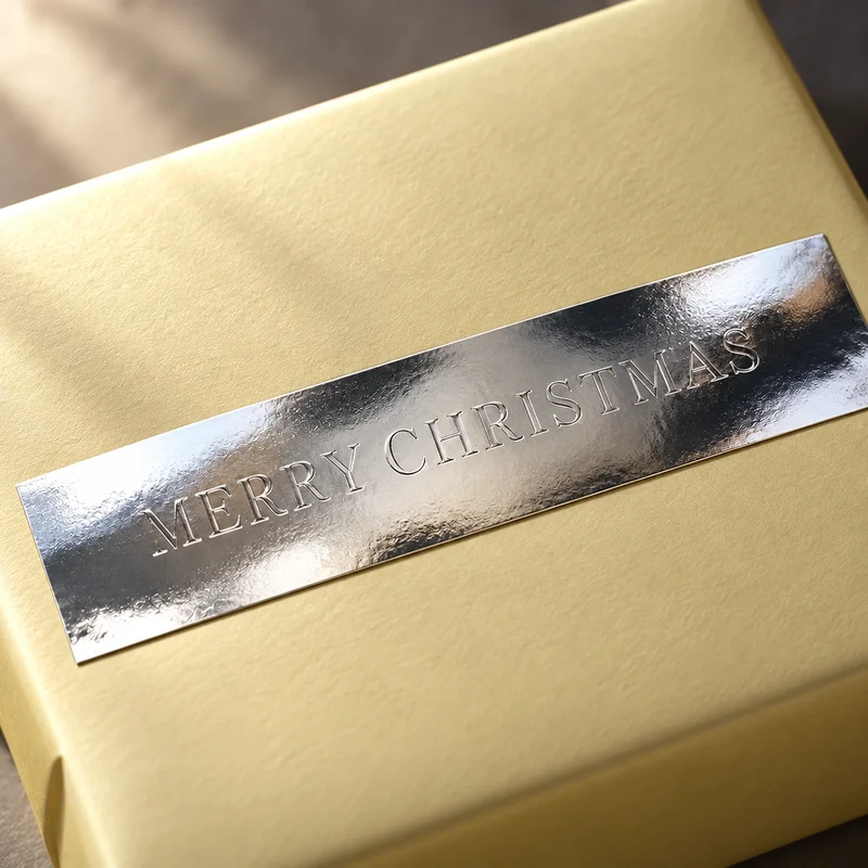 Silberner Embacollage SILVIO MERRY CHRISTMAS Aufkleber mit Blindprägung auf einer goldenen Geschenkbox, festliche Weihnachtsverpackung