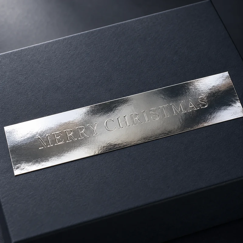Silberfarbener Embacollage SILVIO MERRY CHRISTMAS Aufkleber mit Blindprägung auf einer dunklen Geschenkbox, elegante Weihnachtsverpackung