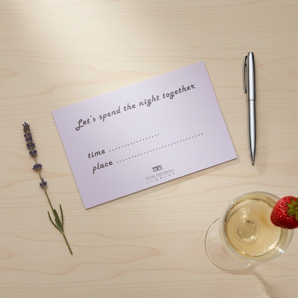 Trixi Gronau LET'S SPEND THE NIGHT TOGETHER notepad in soft lilac, photographed flat on a pale oak desk. With a slim chrome ballpoint pen, a sprig of lavender and a chilled champagne coupe with a strawberry. Playful Hamburg date-night moment. Trixi Gronau