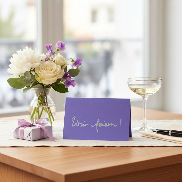 Trixi Gronau WIR FEIERN ! invitation card, B6 landscape in purple-violet pearlescent paper with the phrase Wir feiern ! in flowing light-grey handwritten script centered. On a light-oak desk with a cream linen runner, beside a bouquet of white peonies, cr