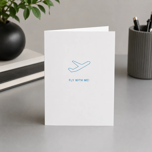 Trixi Gronau FLY WITH ME greeting card for travels, B6 portrait in white paper with a fine turquoise line-art airplane on the upper third, and below FLY WITH ME! in fine spaced turquoise sans-serif capitals. On a light oak desk with a small vase of white 