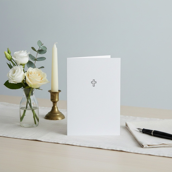 Trixi Gronau Trauerkarte KREUZ, B6 Hochformat in reinem Weiss mit feinem dunkelgrauen Outline-Kreuz in der oberen Mitte. Auf hellem Schreibtisch mit cremefarbenem Leinen-Laeufer, daneben kleines stilles Bouquet aus weisser Eustoma, einer cremefarbenen Ros