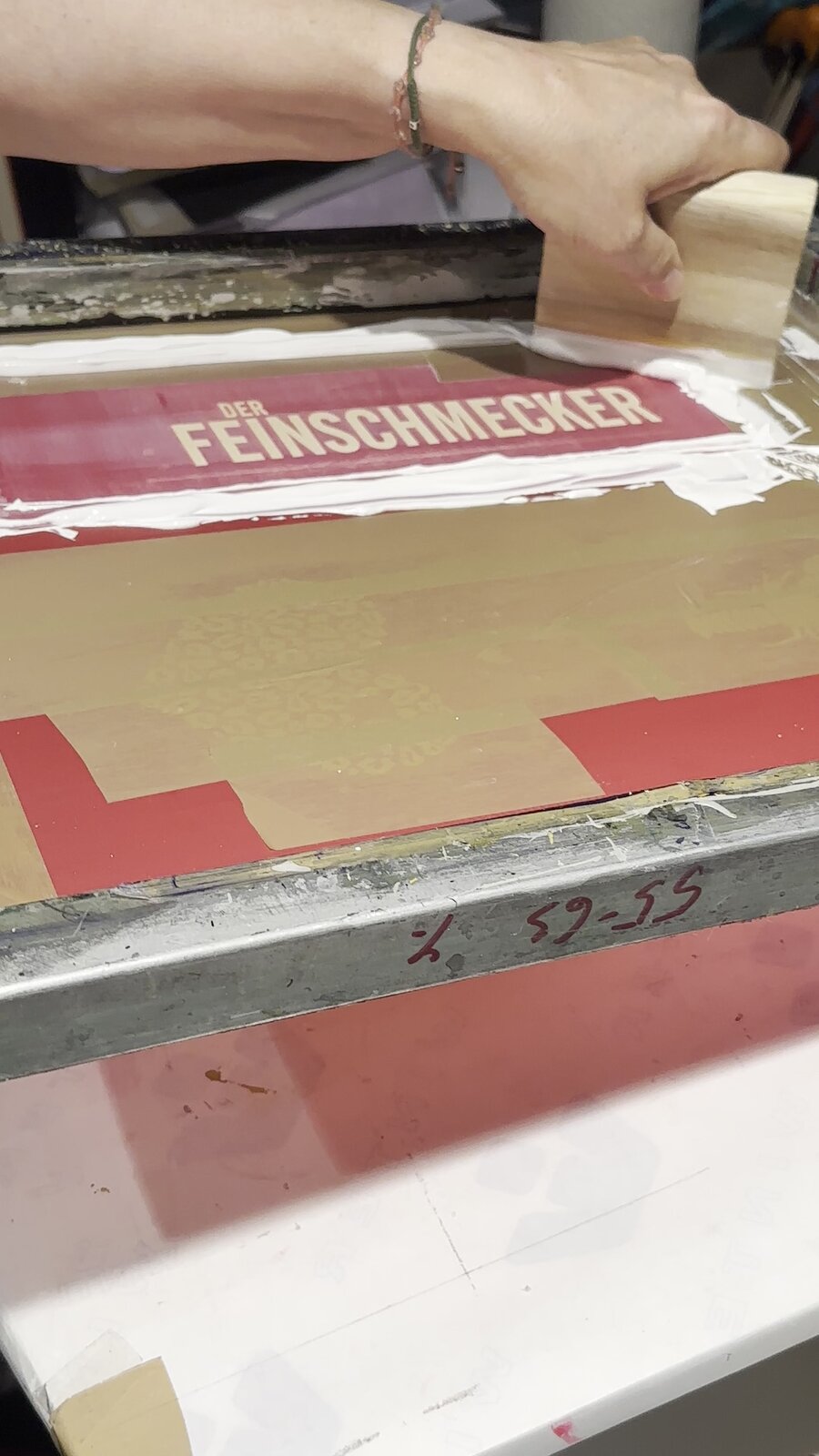 Weiße Siebdruckfarbe wird mit dem Rakel über ein Sieb mit dem Schriftzug Der Feinschmecker gezogen