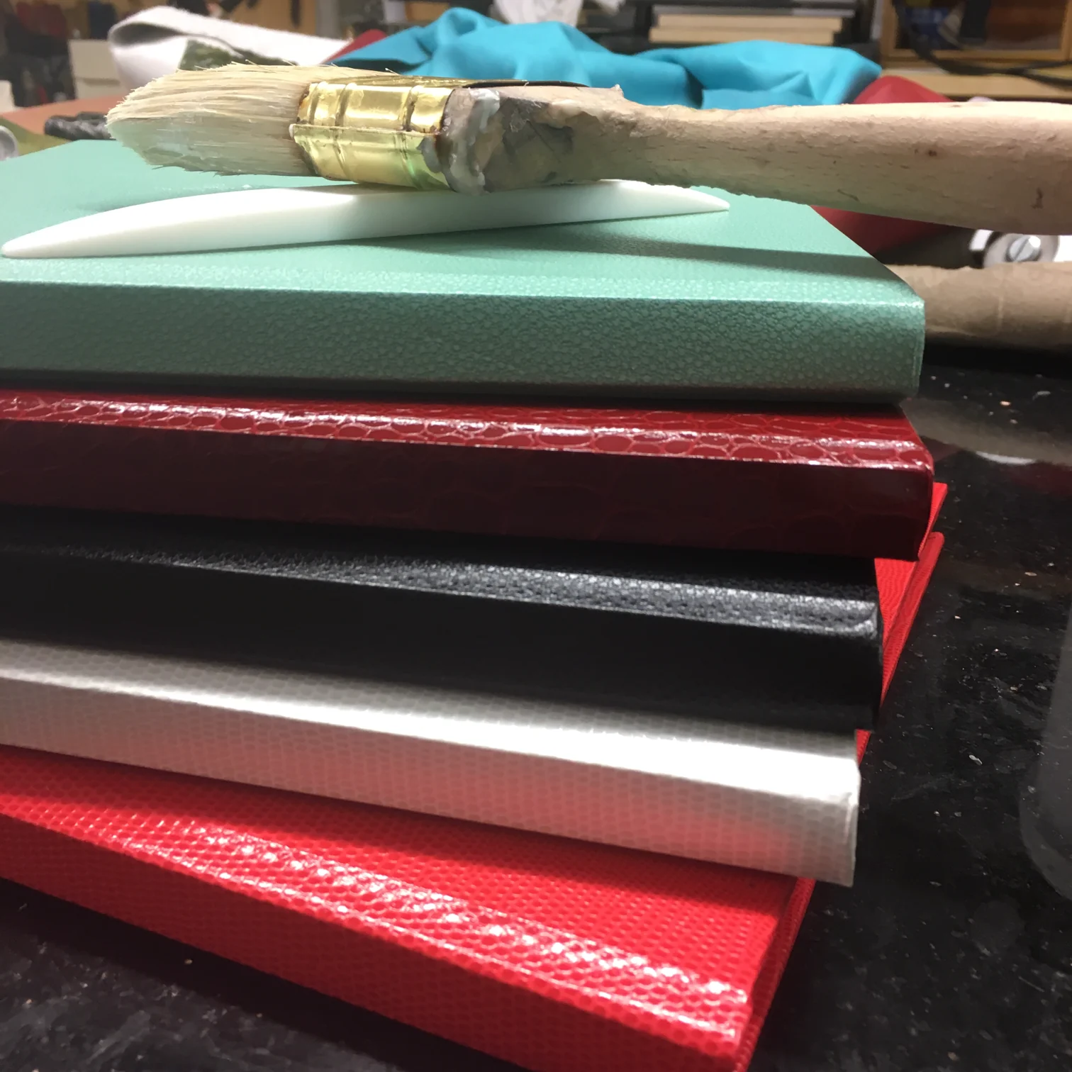 Five finished book covers stacked in mint, dark red, black, white and red, with a bone folder and a glue brush resting on top