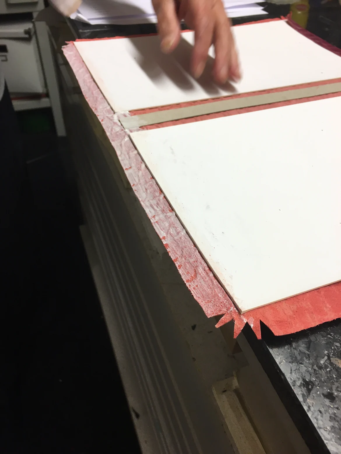 Rotes Bezugsmaterial mit zwei aufgelegten Buchpappen, ringsherum bleibt ein gleichmäßiger Überstand frei, die Ecke ist bereits schräg eingeschnitten