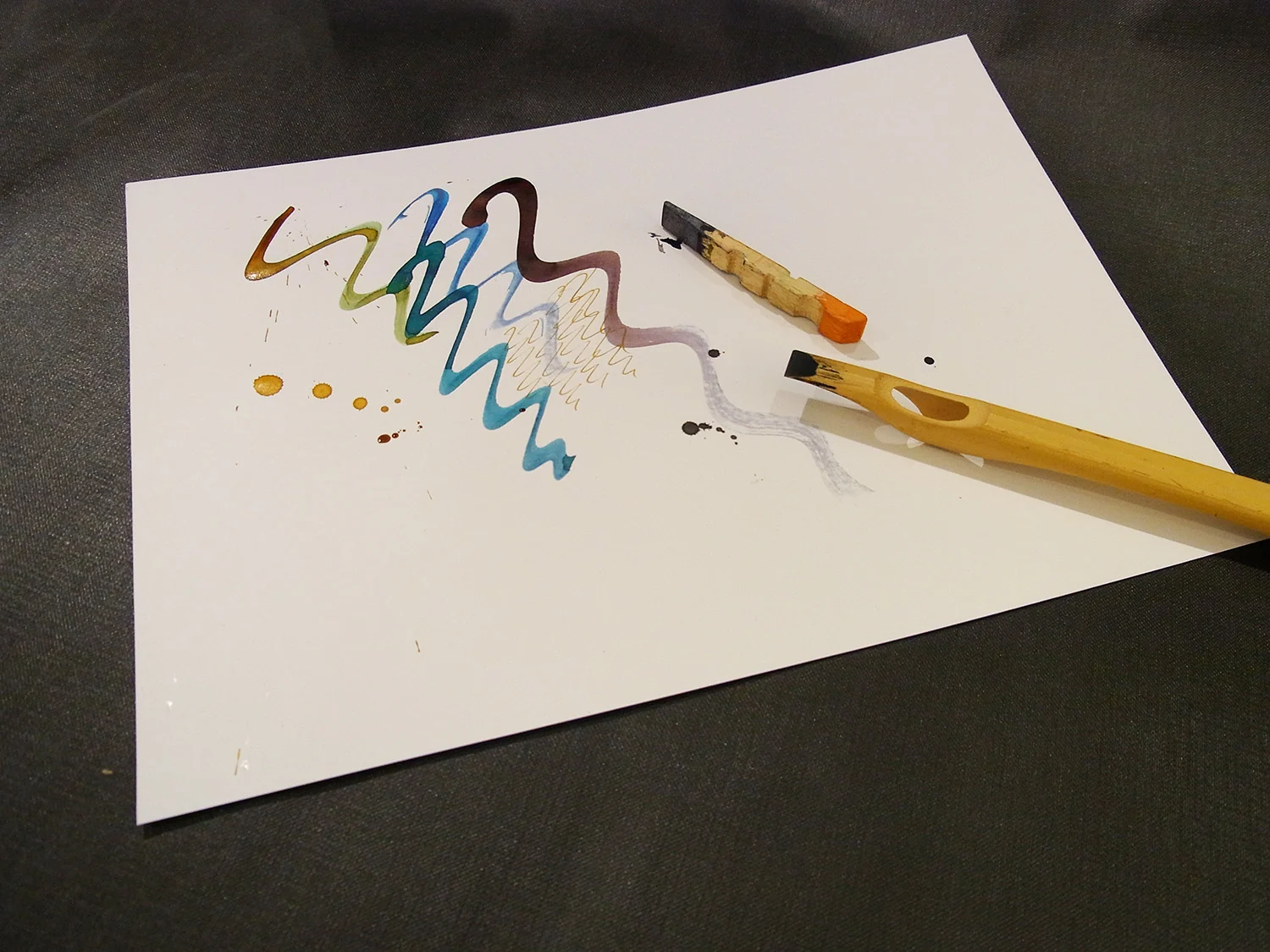 Schriftproben in mehreren Farben auf Papier, daneben eine hölzerne Wäscheklammer und ein Pinsel als Schreibwerkzeug