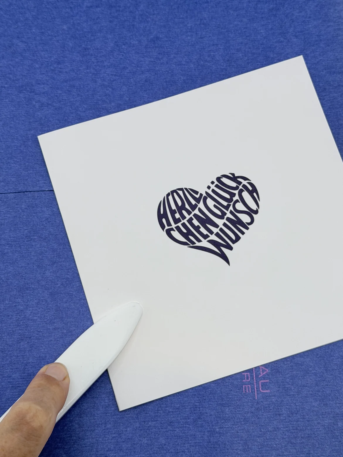 Eine Hand führt ein weißes Falzbein an der Kante einer quadratischen Karte mit dunkelblauem Herz-Schriftzug, der Untergrund ist blaues Papier