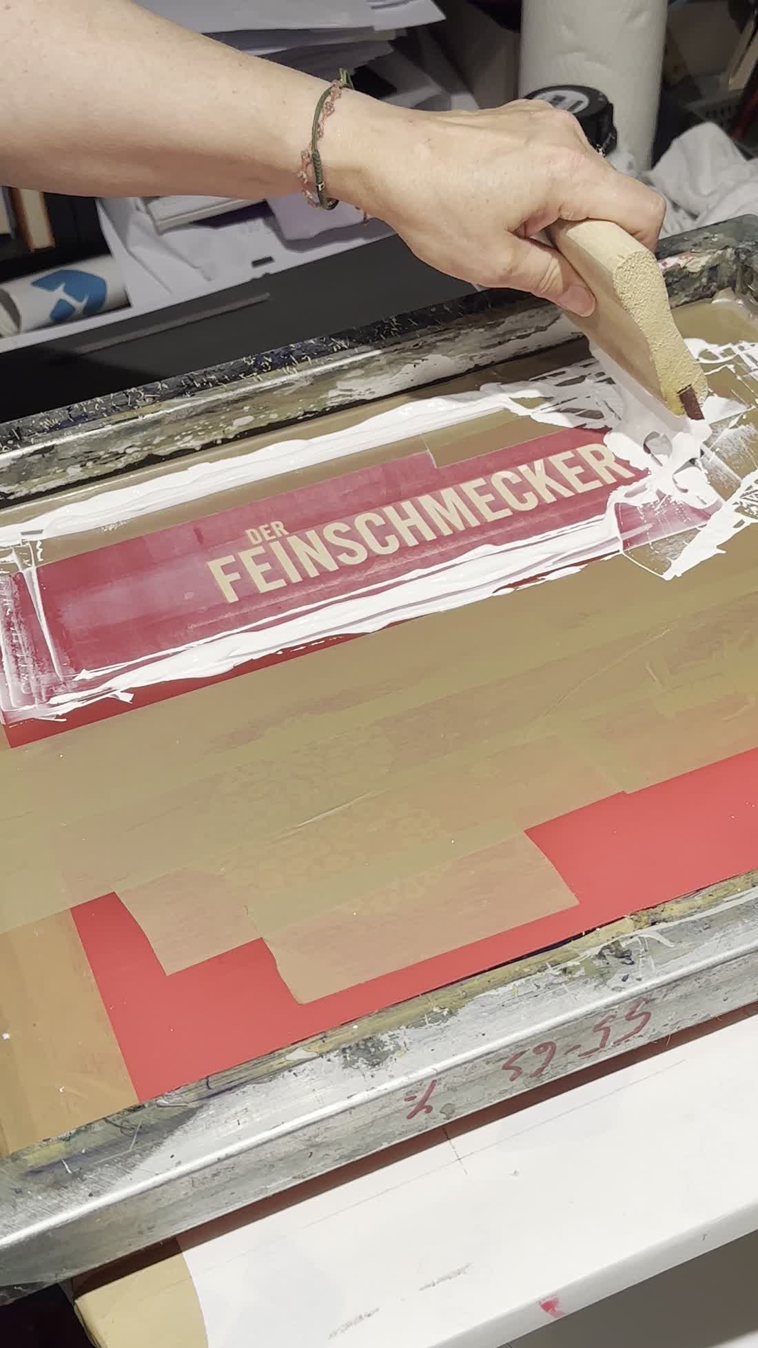 White screen printing ink pulled with a squeegee across a screen carrying the words Der Feinschmecker