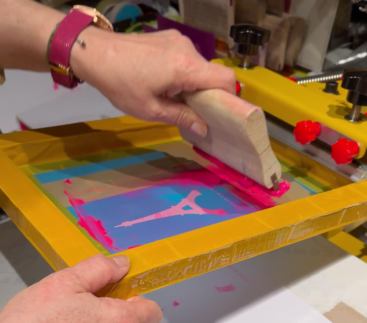 Hand pulling a wooden squeegee with neon pink ink across a yellow framed screen with an Eiffel Tower motif