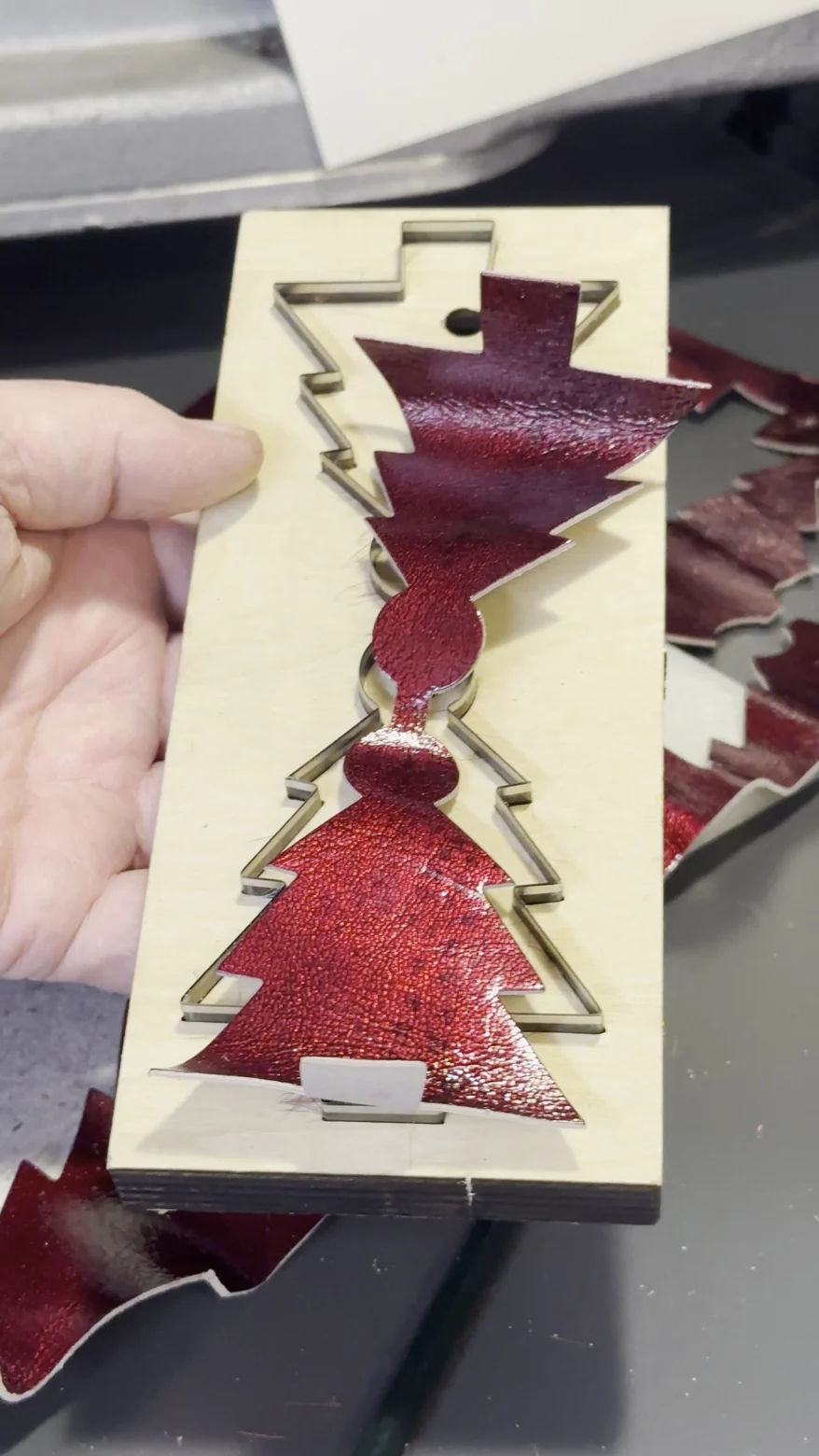 Eine Hand hält eine Stanzform aus Sperrholz mit Tannenbaum-Kontur, darin liegt noch das ausgestanzte rote Glanzleder