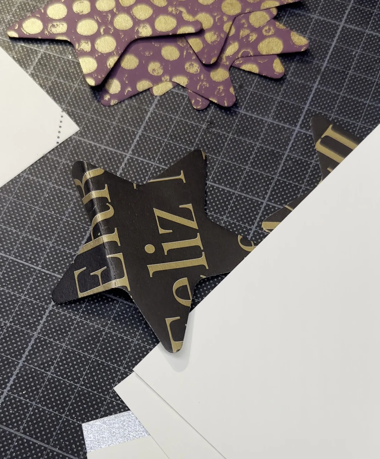 Ausgestanzte Sterne aus verschiedenen Papieren, aubergine mit Goldpunkten und schwarz mit Goldschrift, auf einer Schneidmatte